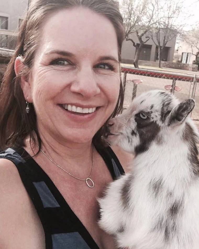 person with long hair, smiling, holding baby goat